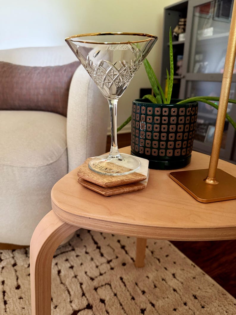 Op de afbeelding: Een helder kristallen martiniglas met een gouden rand, op een houten tafel. Het glas heeft een ruitpatroon en de tekst "The Miller". Twee houten onderzetters met "Sugar" zijn eronder gestapeld. Een plant is zichtbaar.