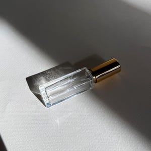 May include: A rectangular clear glass perfume bottle with a metallic gold cap. The bottle features a thin, elongated shape and a subtle, etched script logo on the front surface, reflecting light to create a distinct shadow on the surface below.