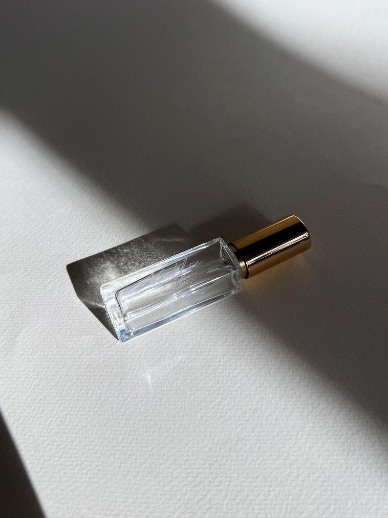 May include: A rectangular clear glass perfume bottle with a metallic gold cap. The bottle features a thin, elongated shape and a subtle, etched script logo on the front surface, reflecting light to create a distinct shadow on the surface below.