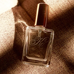 May include: A clear glass perfume bottle with a rectangular shape and a metallic gold cylindrical cap. The front of the bottle features an etched cursive monogram with the letters Ke. The bottle has a transparent pump mechanism inside.