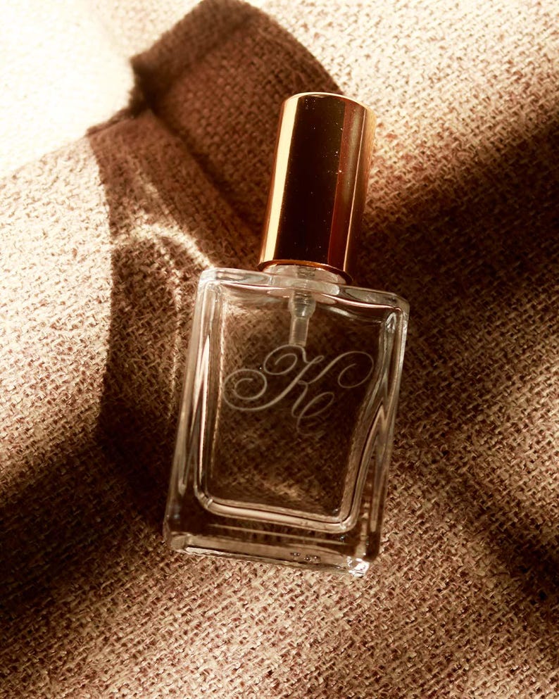 May include: A clear glass perfume bottle with a rectangular shape and a metallic gold cylindrical cap. The front of the bottle features an etched cursive monogram with the letters Ke. The bottle has a transparent pump mechanism inside.
