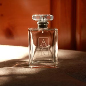 May include: A clear rectangular glass perfume bottle with a faceted crystal-style stopper. The front of the bottle features an etched design with the letters A and K stacked vertically inside a rectangular border. The bottle has a silver spray nozzle.