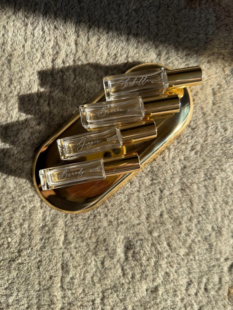 May include: Four rectangular clear glass roller bottles with gold caps, each featuring a white script name printed on the glass. The bottles are arranged diagonally on an oval gold-toned metal tray. The names visible are Isabella, Briana, Ginger, and Brandy.