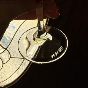 Op de afbeelding: Een helder wijnglas met een lange steel en een brede, cirkelvormige voet. De datum "09.04.2025" is op de voet gegraveerd. Het glas wordt tegen een donkere achtergrond gehouden, met licht dat erdoorheen schijnt.