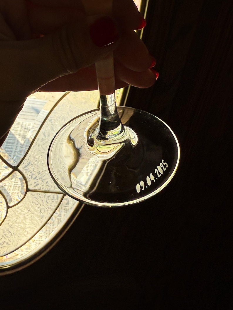 Op de afbeelding: Een helder wijnglas met een lange steel en een brede, cirkelvormige voet. De datum "09.04.2025" is op de voet gegraveerd. Het glas wordt tegen een donkere achtergrond gehouden, met licht dat erdoorheen schijnt.