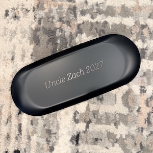 Op de afbeelding: Een matzwarte, ovale opbergdoos met de gegraveerde tekst "Uncle Zach 2027" in het midden van de bovenzijde. De houder heeft gladde, afgeronde randen en een effen, donkere afwerking, wat zorgt voor een strakke en eenvoudige uitstraling.
