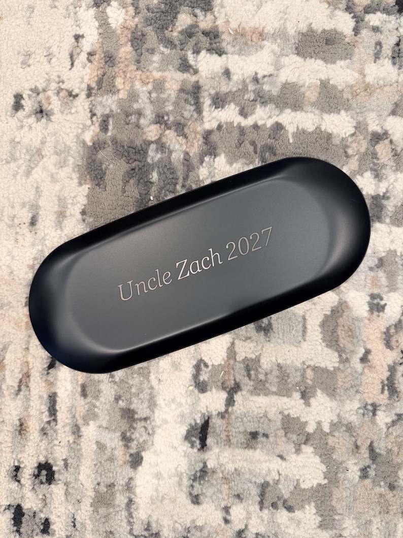 Op de afbeelding: Een matzwarte, ovale opbergdoos met de gegraveerde tekst "Uncle Zach 2027" in het midden van de bovenzijde. De houder heeft gladde, afgeronde randen en een effen, donkere afwerking, wat zorgt voor een strakke en eenvoudige uitstraling.