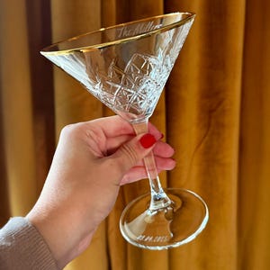 Op de afbeelding: Een helder, ge&euml;tst martiniglas met een gouden rand, gehouden tegen een gouden gordijn. Het glas heeft een ruitpatroon en de inscriptie "The Millers" op de rand en "22.04.2025" op de basis. Een klassiek cocktailglas.