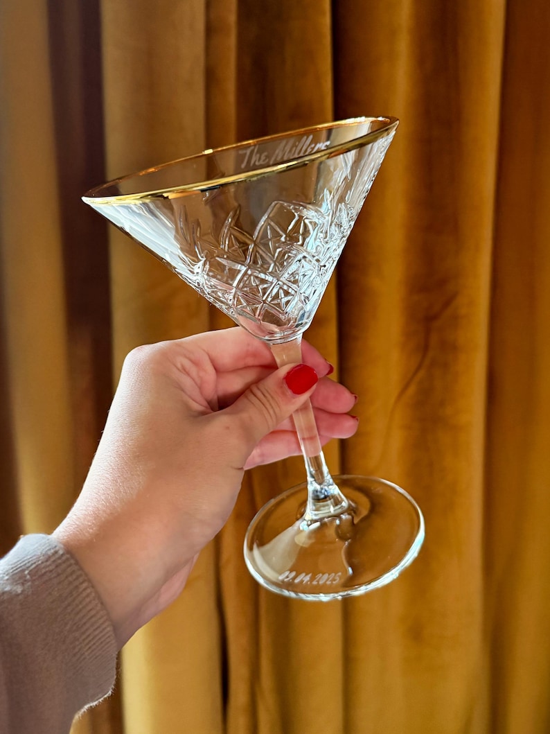Op de afbeelding: Een helder, ge&euml;tst martiniglas met een gouden rand, gehouden tegen een gouden gordijn. Het glas heeft een ruitpatroon en de inscriptie "The Millers" op de rand en "22.04.2025" op de basis. Een klassiek cocktailglas.