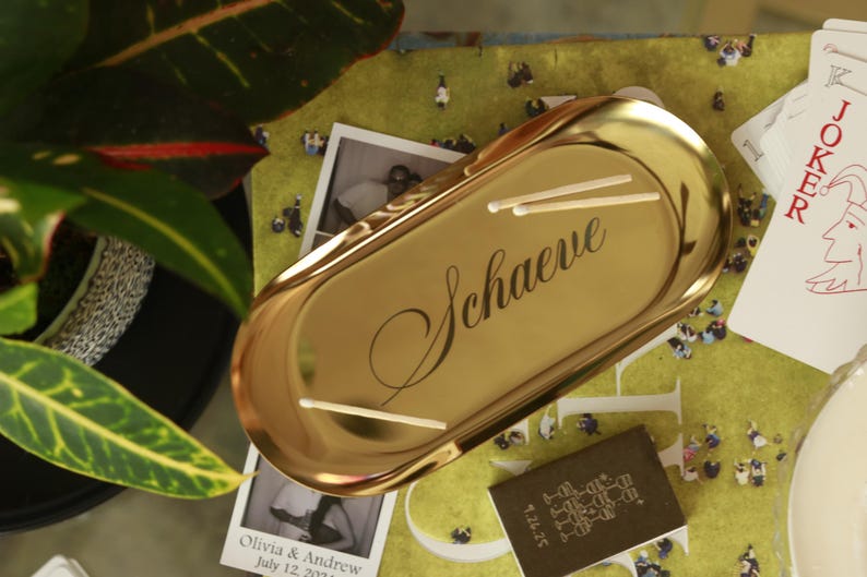 Op de afbeelding: Een ovaalvormig goudkleurig metalen dienblad met een gepolijste afwerking. Het woord "Schaeve" is in zwarte cursieve letters in het midden van het dienblad gegraveerd. Twee dunne, lichtgekleurde houten lucifers liggen op het oppervlak.