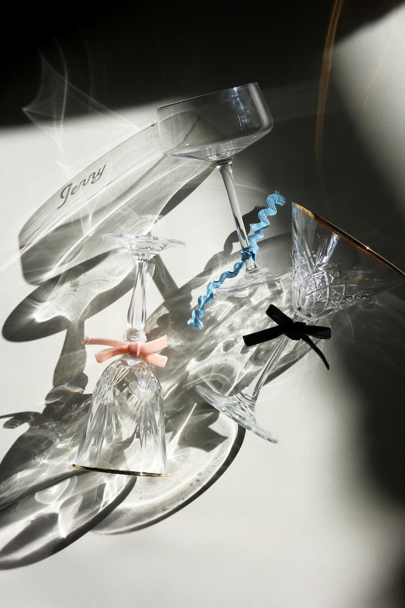 Op de afbeelding: Een assortiment elegant glaswerk, waaronder champagneglazen en een tumbler, gerangschikt op een lichte ondergrond. Sommige glazen zijn versierd met decoratieve linten in roze, blauw en zwart. Een helder acryl stuk met de naam "Jerry" is ook aanwezig.