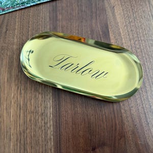 Op de afbeelding: Een ovaalvormig goudkleurig metalen dienblad met een gepolijst, reflecterend oppervlak. Het woord "Tarlow" is gegraveerd in een zwart, elegant cursief lettertype in het midden van het dienblad, dat opstaande randen en een gladde metalen afwerking heeft.