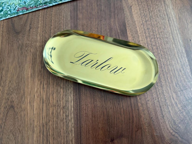 Op de afbeelding: Een ovaalvormig goudkleurig metalen dienblad met een gepolijst, reflecterend oppervlak. Het woord "Tarlow" is gegraveerd in een zwart, elegant cursief lettertype in het midden van het dienblad, dat opstaande randen en een gladde metalen afwerking heeft.