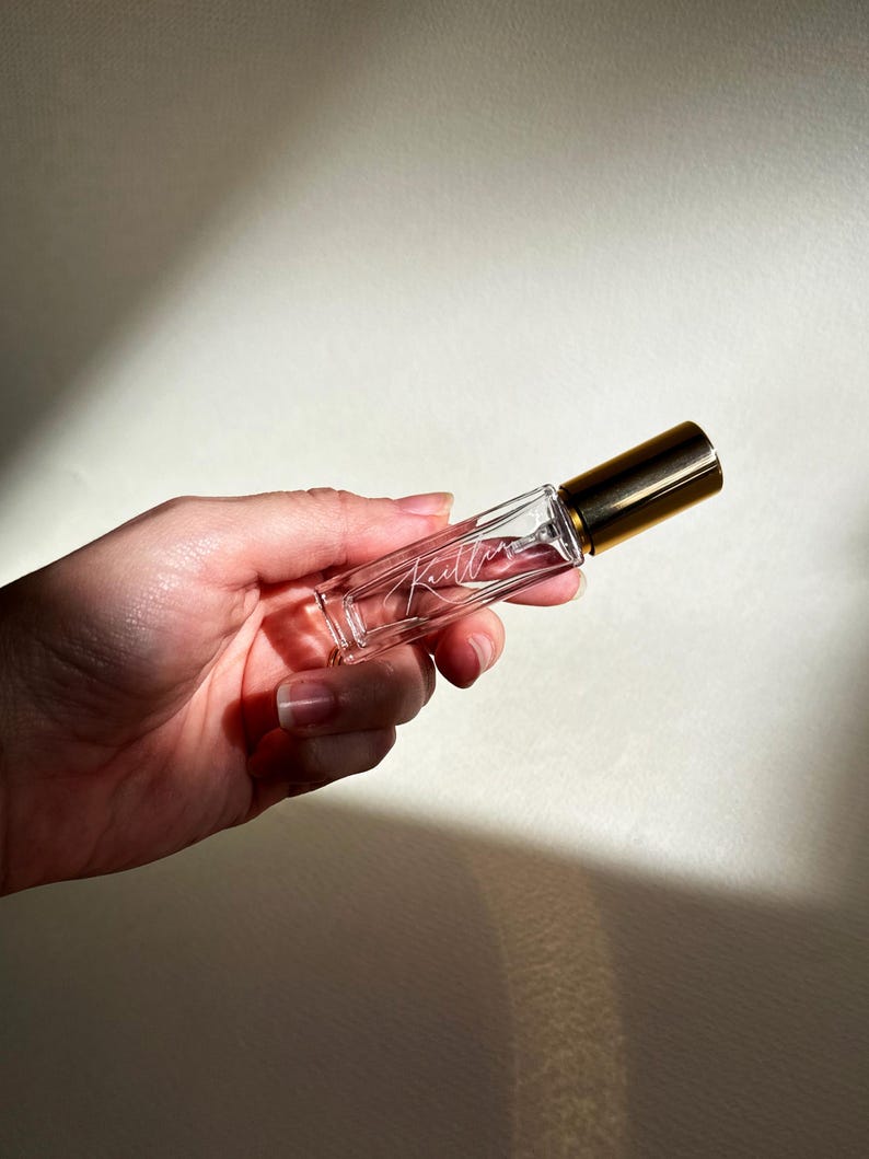 May include: A clear glass perfume bottle with a gold cap, held in a hand. The bottle has the word "Kitten" written in cursive script. The background is a neutral, off-white color.