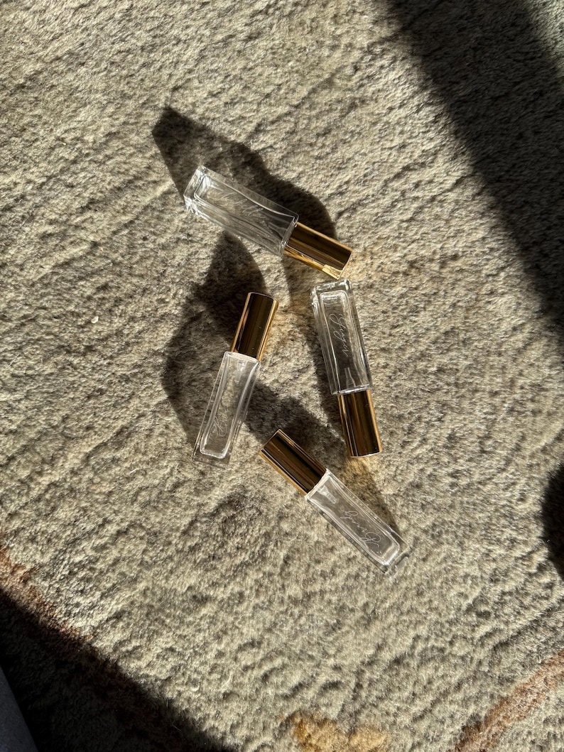 May include: Four rectangular clear glass perfume bottles with gold-colored metal caps. Each bottle features a slender, elongated shape and a transparent body. The bottles are arranged in a scattered formation, reflecting light and casting distinct shadows.