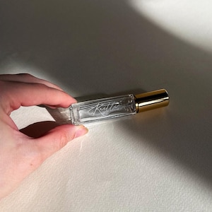 May include: A rectangular clear glass roller bottle with a gold-colored cap. The glass features the white cursive text "Kaitlin" etched or printed on the front surface. The bottle is transparent, revealing the empty interior space within the glass.