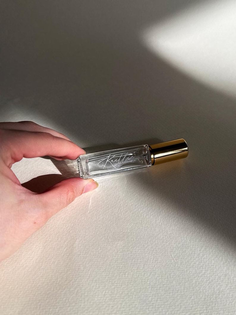 May include: A rectangular clear glass roller bottle with a gold-colored cap. The glass features the white cursive text "Kaitlin" etched or printed on the front surface. The bottle is transparent, revealing the empty interior space within the glass.