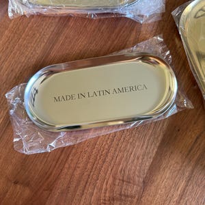Op de afbeelding: Een ovaalvormig goudkleurig metalen dienblad met een gepolijste afwerking, gedeeltelijk verpakt in doorzichtig plastic. Het midden van het dienblad bevat de zwarte tekst "MADE IN LATIN AMERICA" in een strak, schreefloos lettertype.