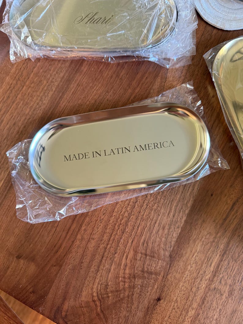 Op de afbeelding: Een ovaalvormig goudkleurig metalen dienblad met een gepolijste afwerking, gedeeltelijk verpakt in doorzichtig plastic. Het midden van het dienblad bevat de zwarte tekst "MADE IN LATIN AMERICA" in een strak, schreefloos lettertype.