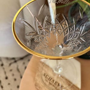 Op de afbeelding: Een helder champagneglas met een gouden rand en een ge&euml;tst bloemmotief. De tekst "The Millers" is op het glas ge&euml;tst. Het glas staat op een houten en witte hars onderzetter met de tekst "SCHAEVE".