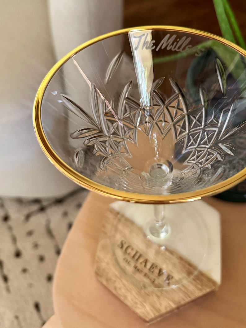 Op de afbeelding: Een helder champagneglas met een gouden rand en een ge&euml;tst bloemmotief. De tekst "The Millers" is op het glas ge&euml;tst. Het glas staat op een houten en witte hars onderzetter met de tekst "SCHAEVE".