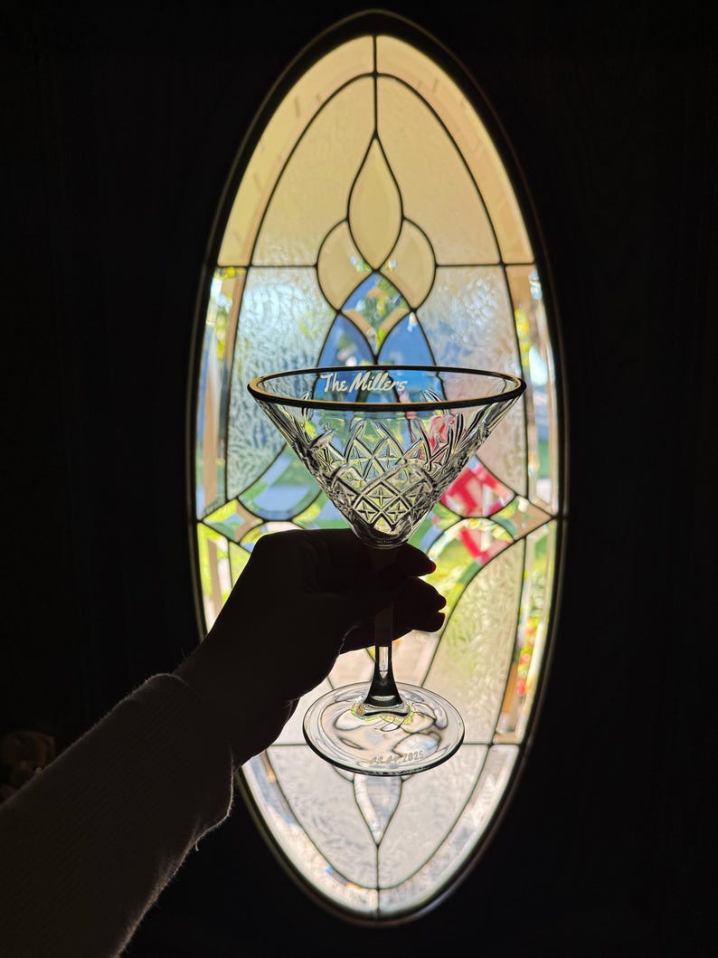 Op de afbeelding: Een helder martiniglas met een gouden rand en ruitpatroon, gehouden voor een glas-in-loodraam. De tekst "The Millers" staat op het glas. Het glas heeft een ronde voet.