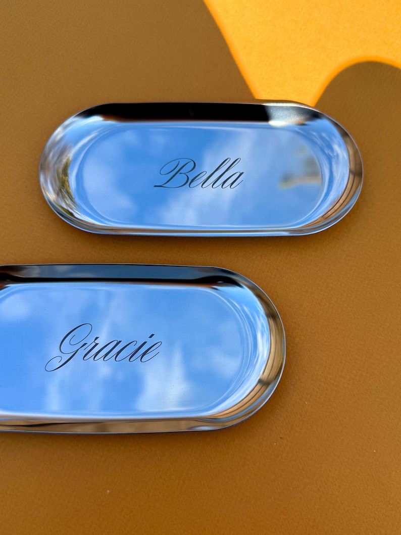 Op de afbeelding: Twee ovale, zilverkleurige metalen dienbladen met een gepolijste, reflecterende afwerking. Het bovenste dienblad is gegraveerd met de naam Bella in cursief schrift, en het onderste dienblad is gegraveerd met de naam Gracie in bijpassend cursief.