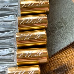 May include: A collection of clear glass spray bottles with shiny gold caps. Each gold cap is engraved with the date 7.11.26 in a cursive script font. The bottles are arranged in a row, showcasing the transparent glass bodies and the metallic finish.