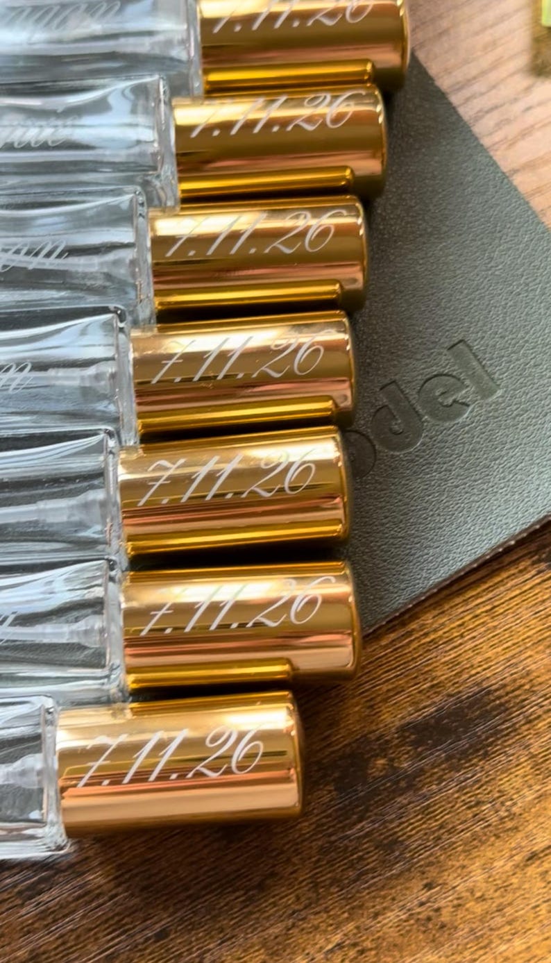 May include: A collection of clear glass spray bottles with shiny gold caps. Each gold cap is engraved with the date 7.11.26 in a cursive script font. The bottles are arranged in a row, showcasing the transparent glass bodies and the metallic finish.