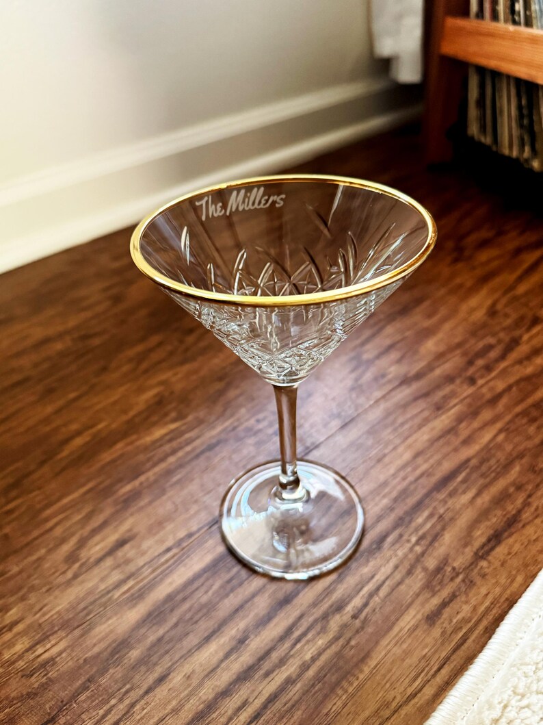Op de afbeelding: Een helder martiniglas met een gouden rand en een ge&euml;tst diamantpatroon. De tekst "The Millers" is op de rand ge&euml;tst. Het glas staat op een houten oppervlak.