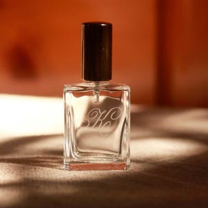 May include: A clear glass perfume bottle with a rectangular shape and a dark brown cylindrical cap. The front of the bottle features an etched script monogram consisting of the letters K and c intertwined in a decorative cursive font style.