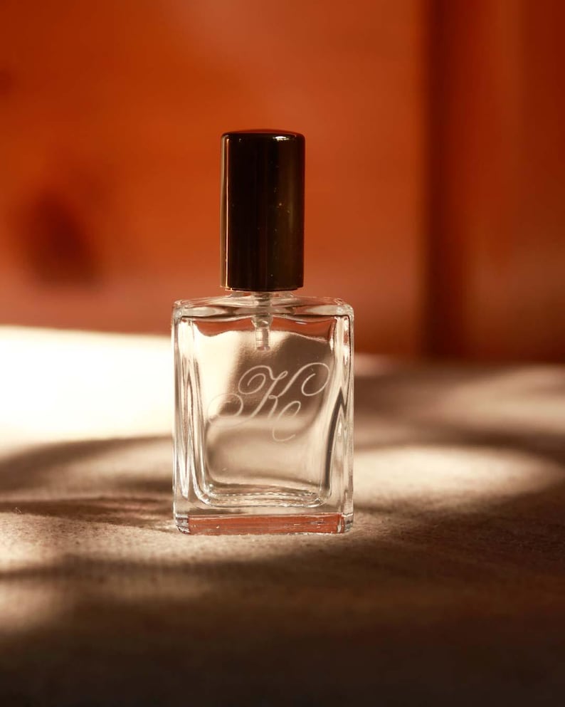 May include: A clear glass perfume bottle with a rectangular shape and a dark brown cylindrical cap. The front of the bottle features an etched script monogram consisting of the letters K and c intertwined in a decorative cursive font style.