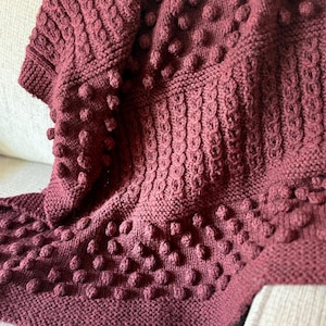 May include: A burgundy knitted blanket with a textured pattern and bobble details. The blanket is folded over a light-colored surface, showcasing the intricate knit design and rich color. The bobbles add a tactile element to the cozy throw.