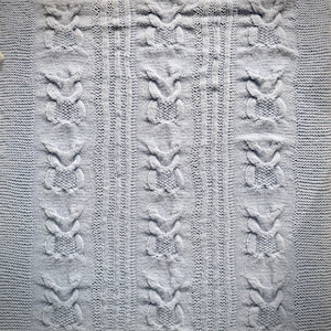 May include: A light blue knitted blanket featuring a repeating cable knit pattern of stylized owl figures arranged in three vertical columns, separated by textured vertical stripes, with a garter stitch border framing the entire rectangular piece.
