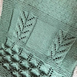 May include: Close-up of a knitted blanket in a muted teal color. The blanket features intricate leaf patterns and textured stitches. The design includes a central panel with leaf motifs and a border with a diamond-like pattern.