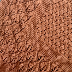 May include: Close-up of a knitted blanket in a warm, earthy brown colour. The blanket features a detailed leaf pattern along the edges and a textured stitch design. The soft, cosy texture is visible throughout the blanket.