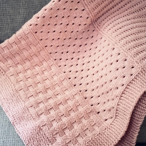 May include: A knitted blanket in a soft rose color. The blanket features various textured patterns, including a diamond stitch and a dotted design. The edges are neatly finished with a ribbed border, creating a cozy and inviting appearance. The blanket is folded, showcasing its intricate details.