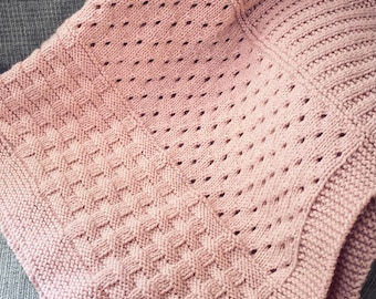 May include: A knitted blanket in a soft rose color. The blanket features various textured patterns, including a diamond stitch and a dotted design. The edges are neatly finished with a ribbed border, creating a cozy and inviting appearance. The blanket is folded, showcasing its intricate details.