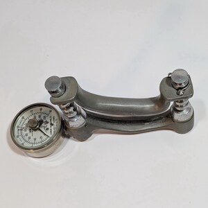 May include: A vintage silver-toned hand dynamometer with a circular gauge. The gauge displays measurements in a circular format. The dynamometer has a curved handle and adjustable settings. The device is designed for measuring grip strength.