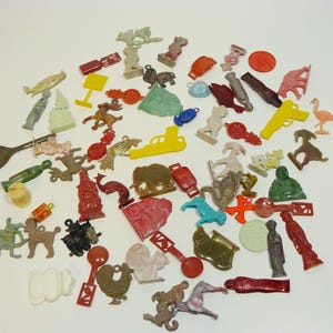 May include: A collection of small, colorful plastic charms and figurines. The assortment includes animals, people, and various objects in shades of red, yellow, green, blue, and brown. A vintage spoon is also present. The charms are scattered on a white surface.