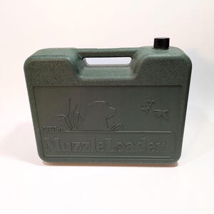 May include: A dark green plastic container with a handle, a black cap, and the text "MuzzleLeader". The container has a textured surface and features a silhouette of a dog, reeds, and flying ducks.