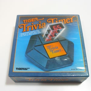 May include: A blue and orange box for the Tiger Trivia Timer game. The box features a red digital timer with the text "TIGER Trivia Timer" on the front. The box also includes the text "Programmable countdown timer, Handicaps to equalize game play, Use with any Trivia question game."