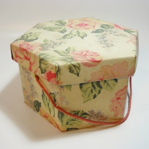 May include: A hexagonal box with a floral pattern in shades of pink, green, and beige. The box has a red rope handle.