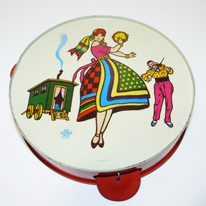 May include: A vintage metal toy tambourine with a colorful illustration of a woman in a folk costume playing a tambourine, a man playing a violin, and a green wagon. The tambourine has a red rim and red handles.