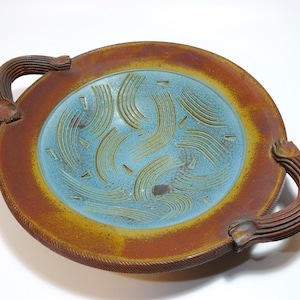 May include: A handcrafted ceramic serving dish with a round shape and two handles. The dish features a blue center with etched wave patterns, surrounded by a brown and yellow rim. The handles are brown and textured.