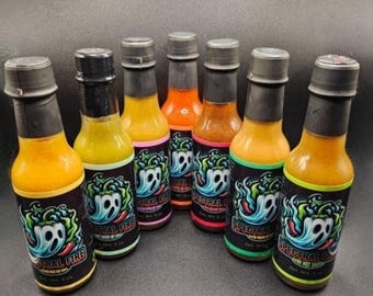 Op de afbeelding: Zeven flessen hete saus met zwarte doppen en etiketten. De etiketten tonen een spookafbeelding en de tekst "SPECTRAL FIRE". De flessen bevatten oranje en gele hete sauzen.