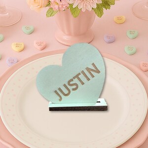 May include: A romantic table setting with a floral centerpiece, plates, and glassware. Heart-shaped candies and signs with the text "LOVE YOU", "KISS ME", and "JUSTIN" are scattered around the table. The color palette is soft pinks and pastels.