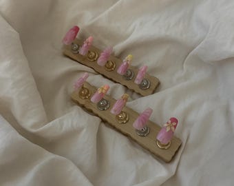 May include: Two wooden nail display stands, each holding six artificial nails. The nails feature various designs, including pink gradients, floral accents, and glitter. The stands are on a white, textured surface.