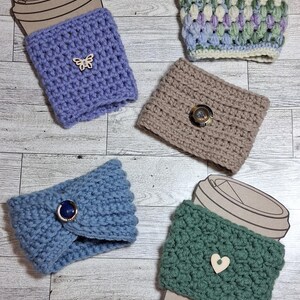 May include: Four crochet cup sleeves in purple, beige, blue, and green. The purple sleeve has a wooden butterfly button, the beige has a dark button, the blue has a dark blue button, the green has a wooden heart button, and the multi-colored one has a green button.