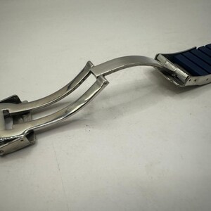 May include: A close-up of a watch strap with a silver clasp. The strap is dark blue and segmented. The clasp is open, showing the mechanism. The background is a plain, light surface.